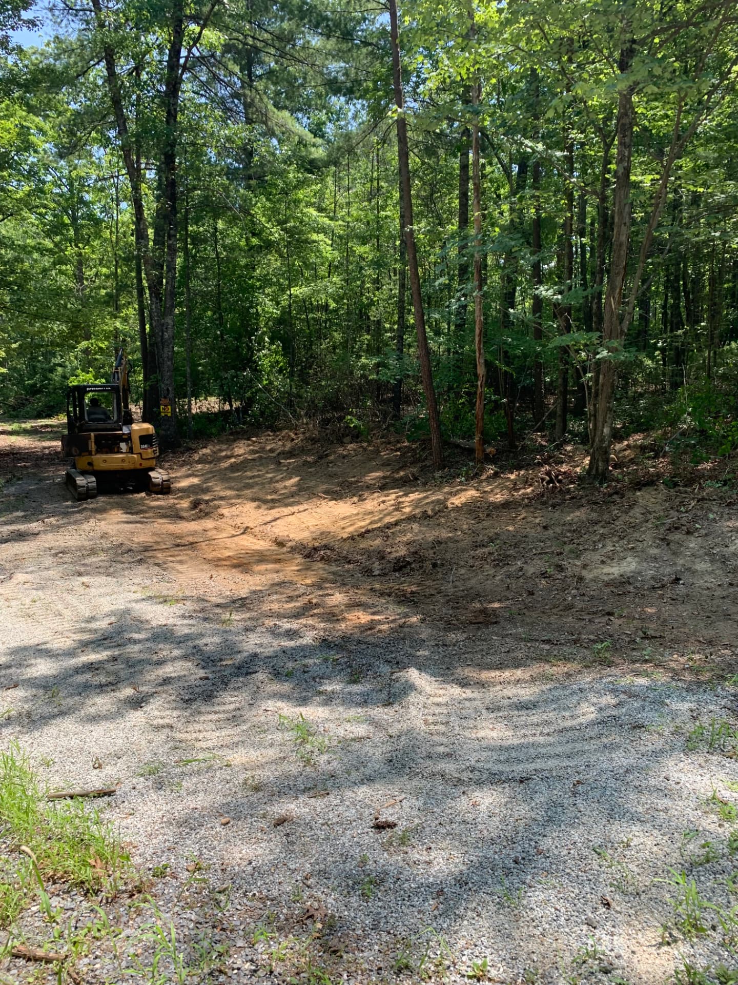 Mini excavator grading a cleared gravel driveway along a tree line